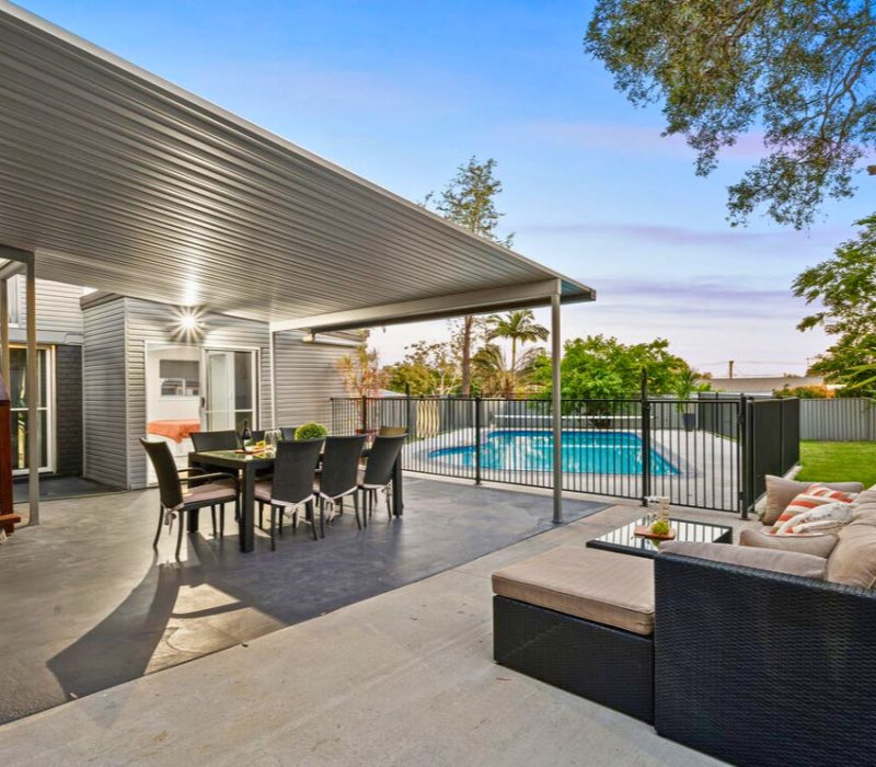 Outdoor dining area under cover beside pool with garden backdrop